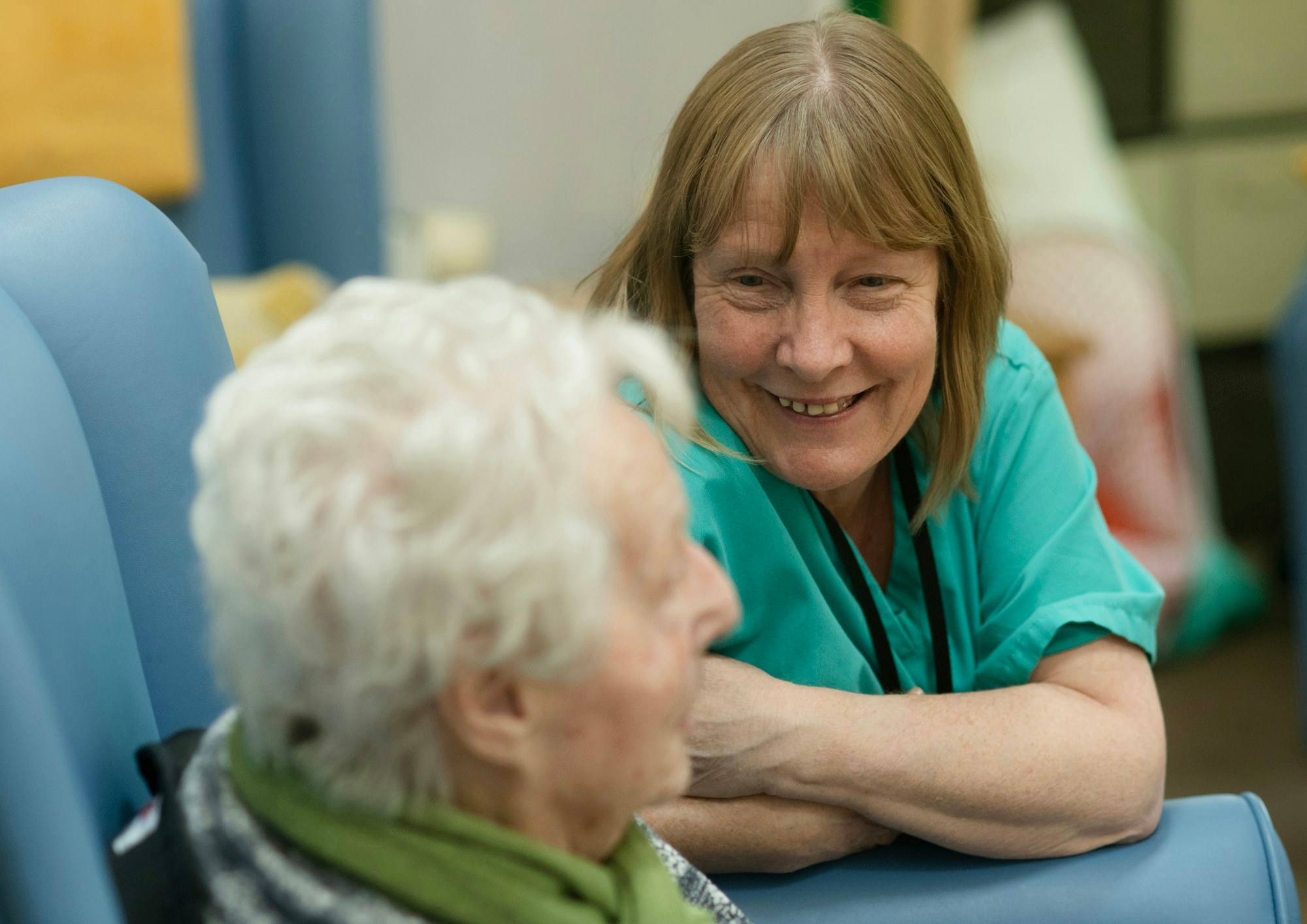 A smiling caregiver in green scrubs talking with an older person