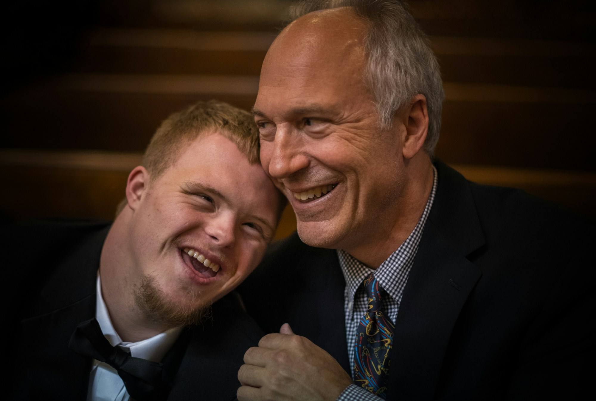 A young man with Down syndrome and an older man in suits, smiling cheek to cheek
