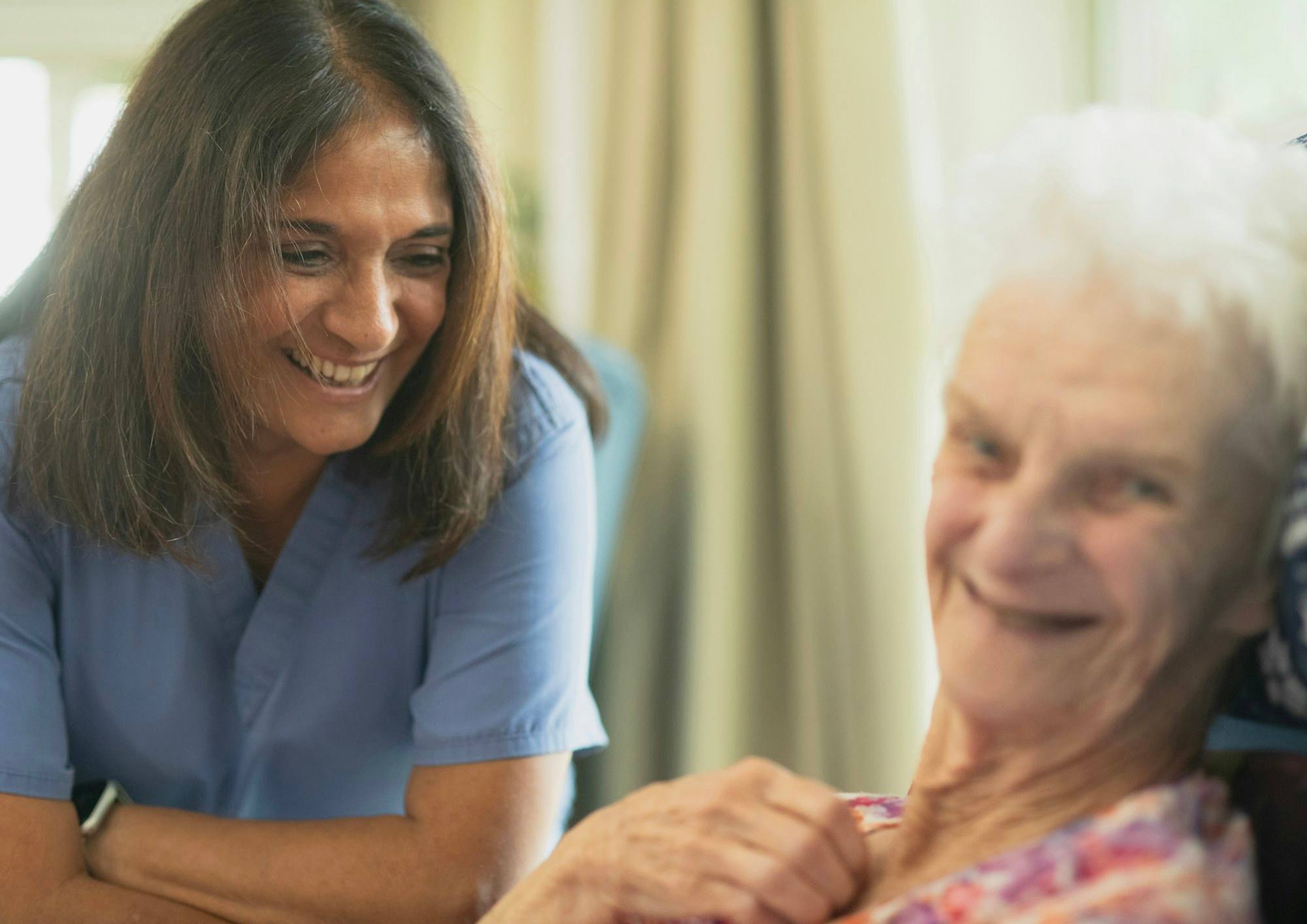 A caregiver in a blue uniform laughing with an older woman