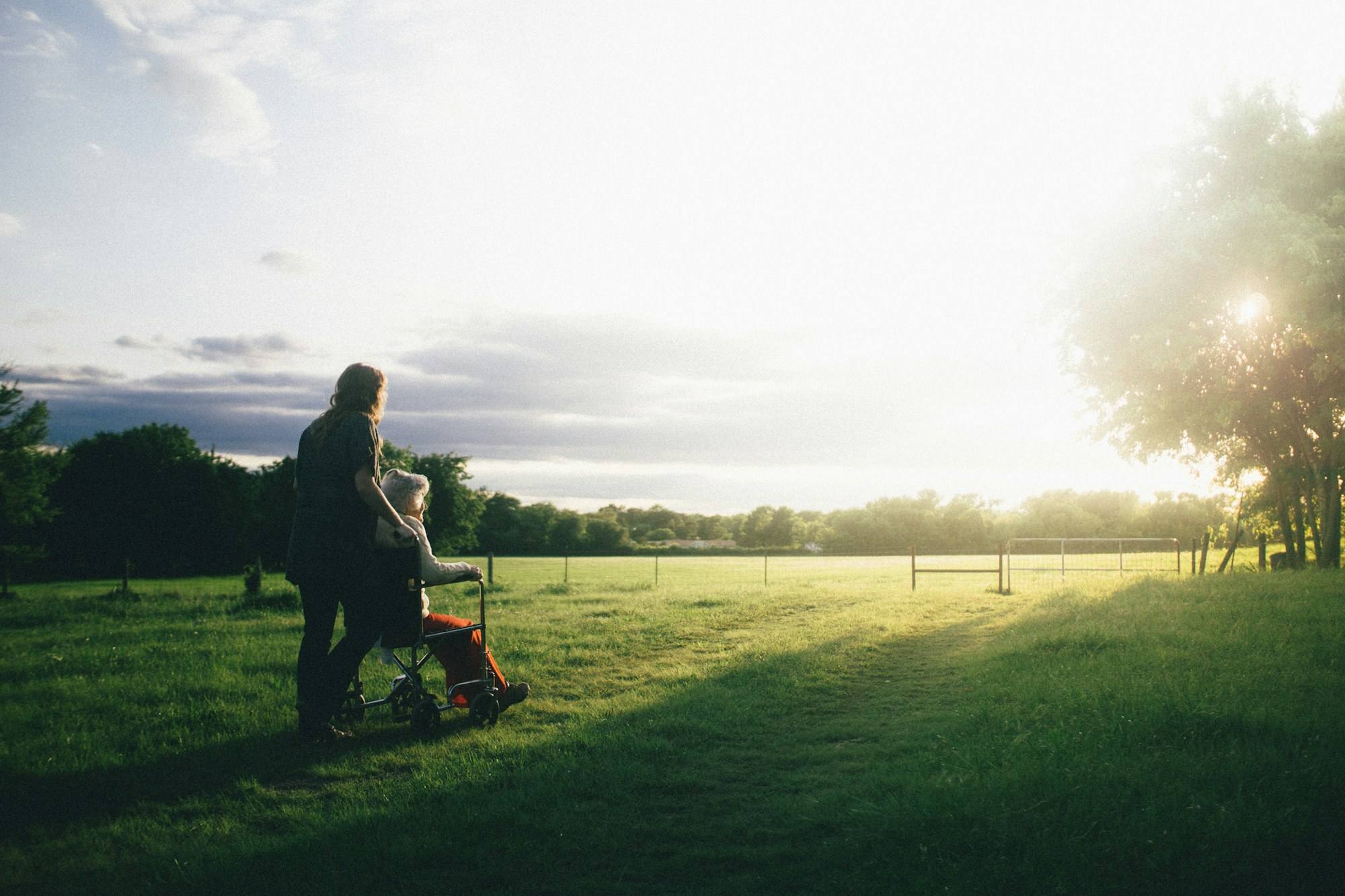 A woman pushing a person in a wheelchair across a field at sunset