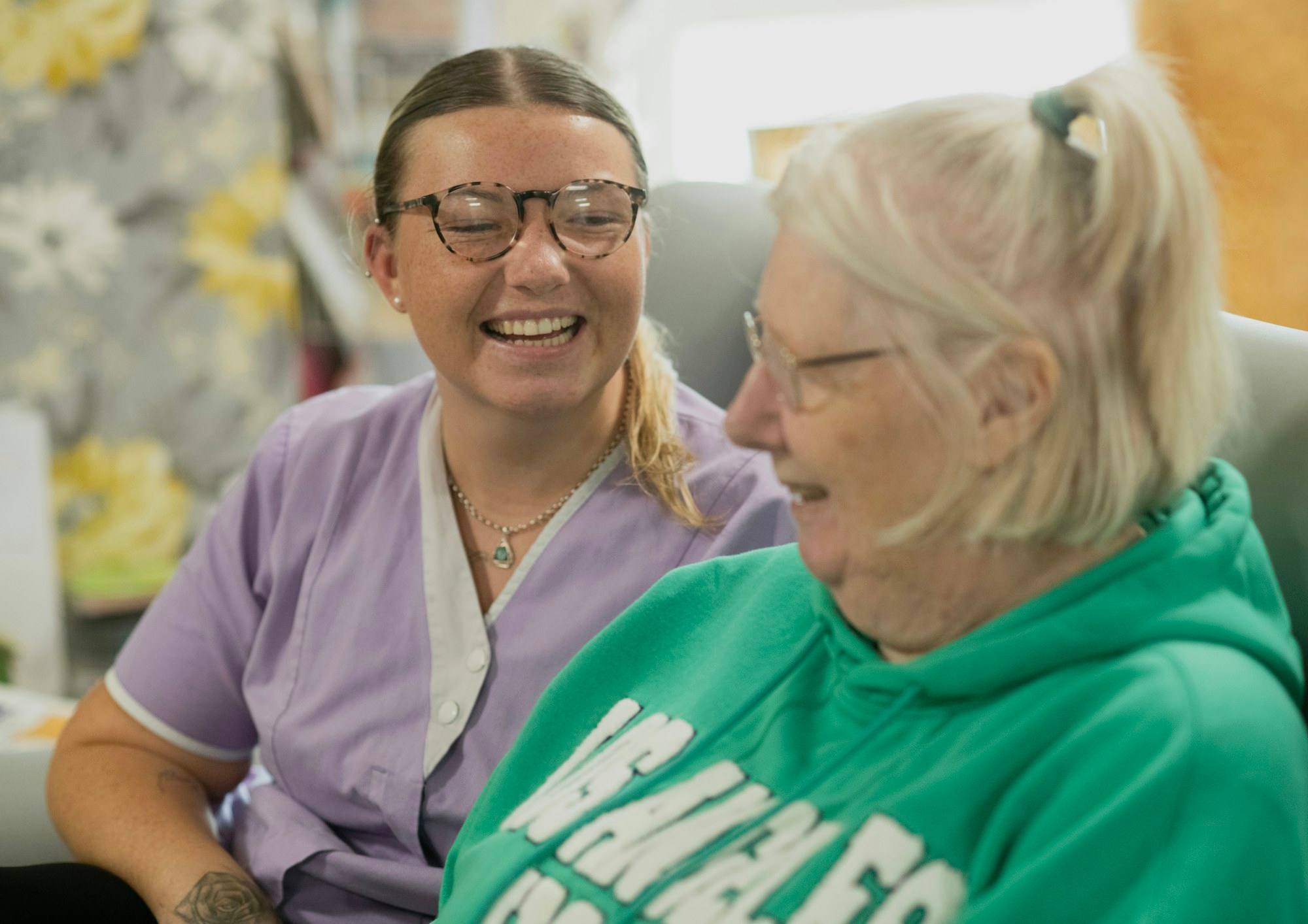 A caregiver and an older woman in a green hoodie laughing together