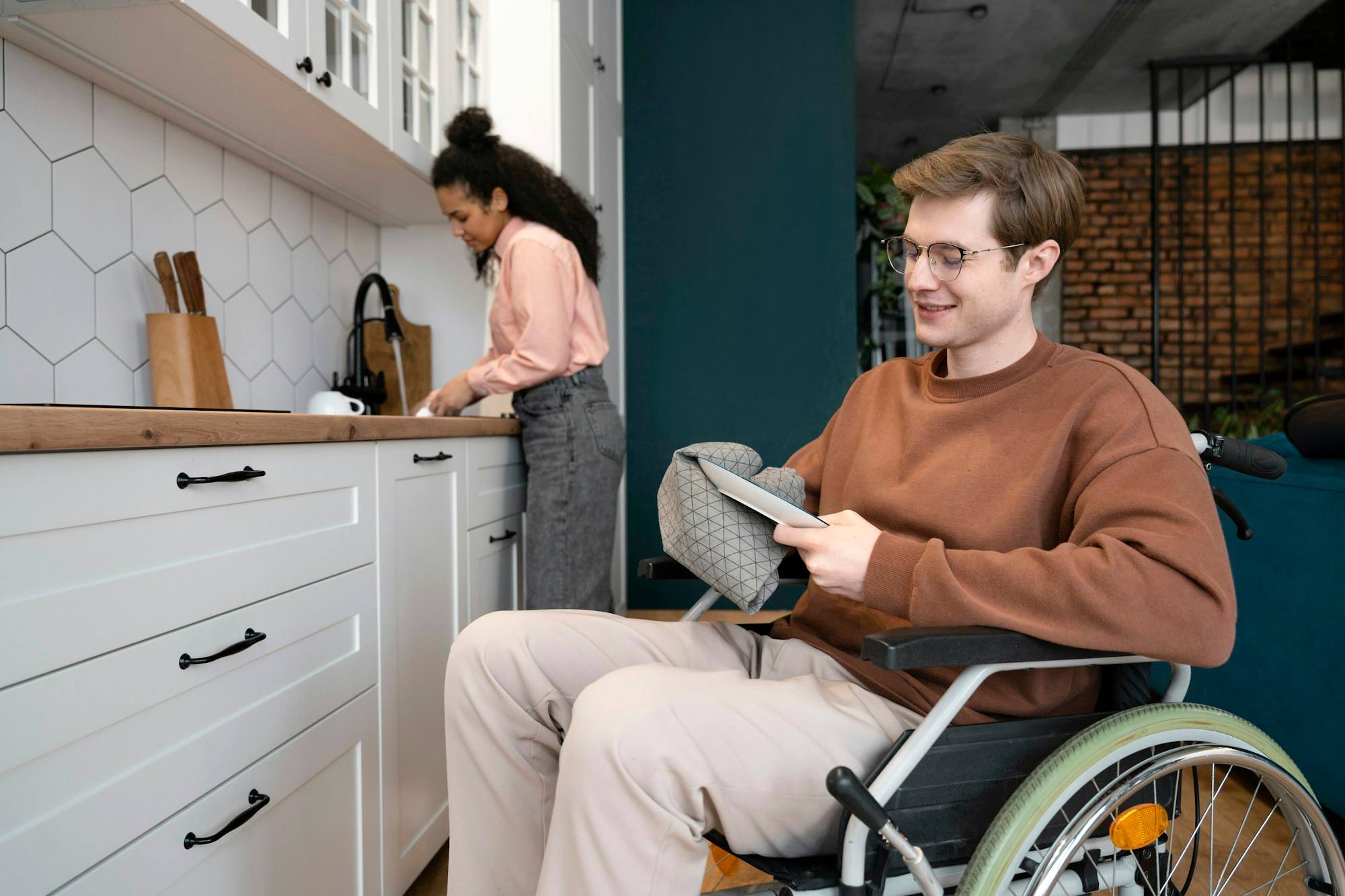 A young man in a wheelchair reading in a kitchen while a woman cooks